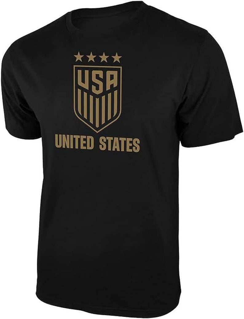May include: Black t-shirt with a gold graphic of the United States soccer team logo. The logo features five stars above the letters "USA" and a shield with vertical stripes. The text "UNITED STATES" is printed below the logo.
