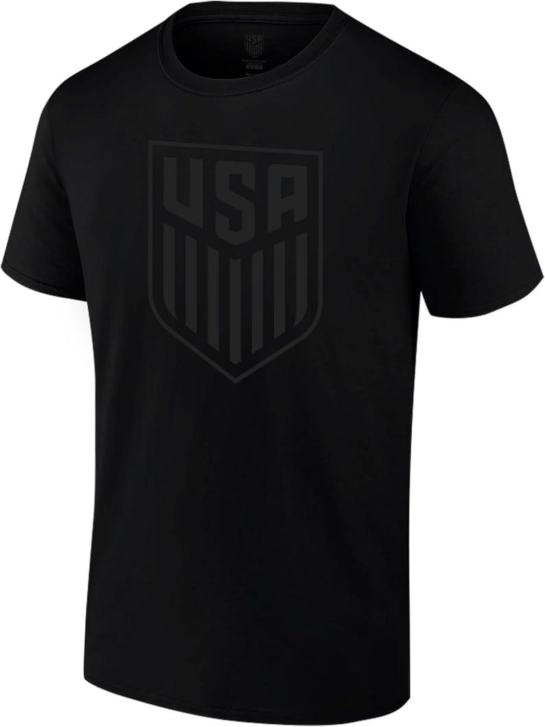 May include: Black crew neck t-shirt featuring a tonal graphic of the letters "USA" inside a shield. The shirt is made of a soft material and has short sleeves. The design is centered on the chest.
