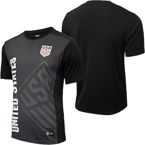 Puede incluir: Camiseta de f&uacute;tbol negra de manga corta con un dise&ntilde;o gr&aacute;fico gris y blanco con las palabras "United States" y "USA".