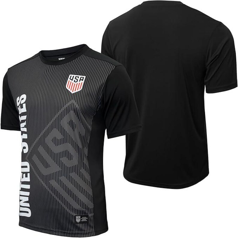 Puede incluir: Camiseta de f&uacute;tbol negra de manga corta con un dise&ntilde;o gr&aacute;fico gris y blanco con las palabras "United States" y "USA".