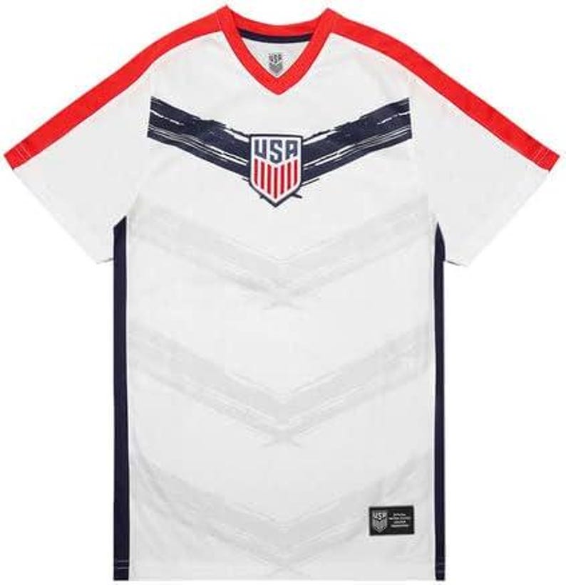 May include: White athletic jersey with red trim on the shoulders and neckline. The jersey features a navy blue stripe across the chest and a USA crest. The jersey has a V-neck and short sleeves. The bottom right corner has a small black tag.