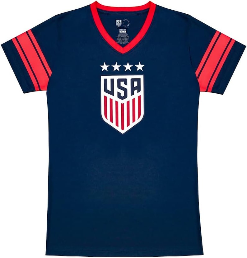 May include: A navy blue t-shirt with red and white stripes on the sleeves. The shirt has a v-neck and features a white shield with the word "USA" and five stars above it.