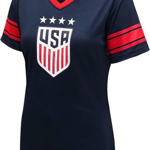 K&ouml;nnte beinhalten: Ein marineblaues T-Shirt mit kurzen &Auml;rmeln und roten und wei&szlig;en Streifen an den &Auml;rmeln. Das Shirt zeigt ein wei&szlig;es Schild mit roten Streifen und dem Wort "USA" in wei&szlig;en Buchstaben. F&uuml;nf wei&szlig;e Sterne befinden sich &uuml;ber dem Schild.