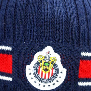 May include: Blue knitted beanie with a red and white striped band and a white embroidered logo of a football team. The logo features a crest with a shield, a crown, and the text "Club Deportivo Guadalajara S.A. de C.V."