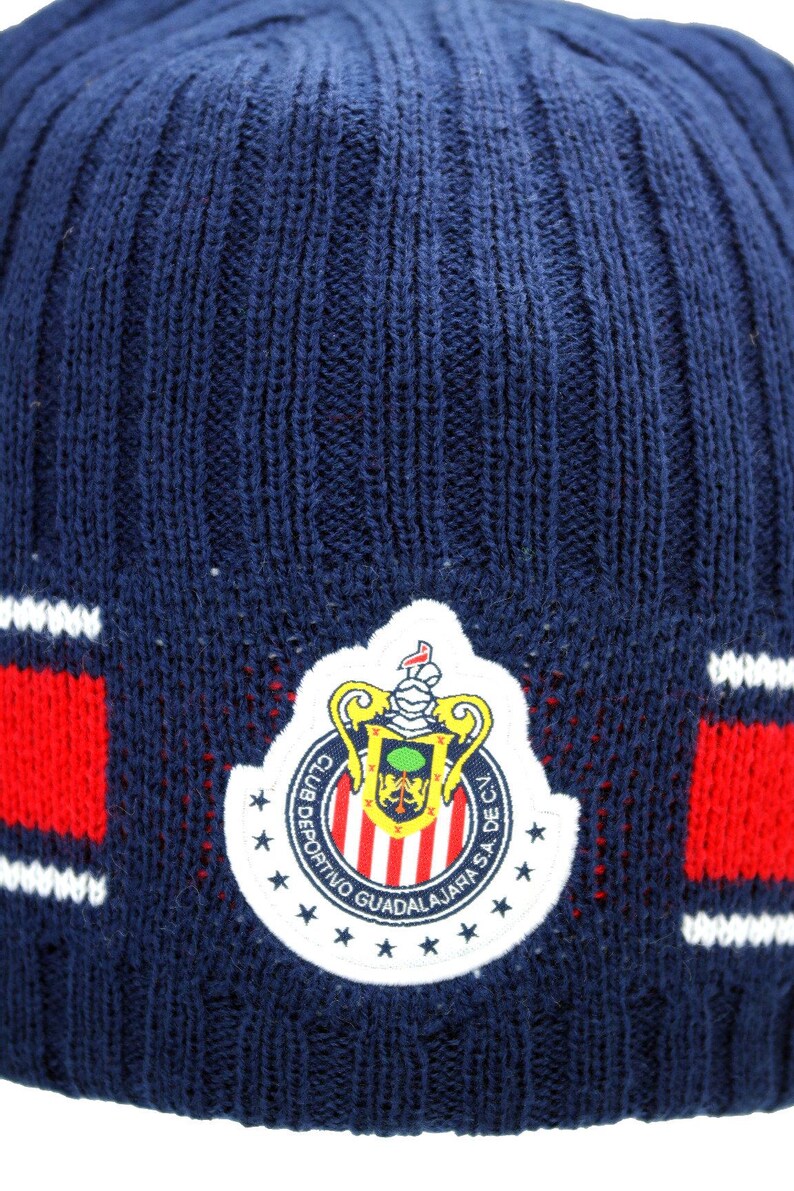 May include: Blue knitted beanie with a red and white striped band and a white embroidered logo of a football team. The logo features a crest with a shield, a crown, and the text "Club Deportivo Guadalajara S.A. de C.V."