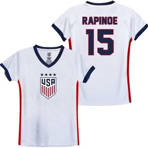 May include: White soccer jersey with red and blue trim. The front of the jersey has a USA crest with stars and stripes. The back of the jersey has the name "RAPINOE" and the number "15" in blue with red outlines.