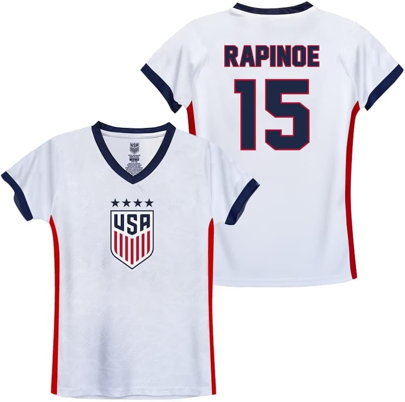 May include: White soccer jersey with red and blue trim. The front of the jersey has a USA crest with stars and stripes. The back of the jersey has the name "RAPINOE" and the number "15" in blue with red outlines.