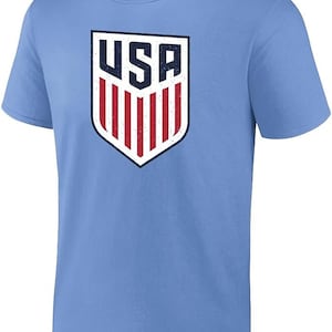 Pode incluir: Camiseta azul clara com o logotipo do escudo dos EUA desgastado. O logotipo inclui as letras "USA" em azul marinho e listras vermelhas sobre um fundo branco. A camiseta tem gola redonda e mangas curtas.