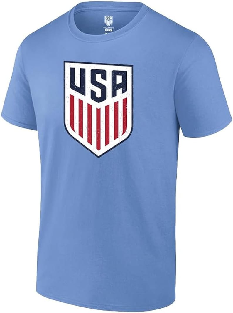 Pode incluir: Camiseta azul clara com o logotipo do escudo dos EUA desgastado. O logotipo inclui as letras "USA" em azul marinho e listras vermelhas sobre um fundo branco. A camiseta tem gola redonda e mangas curtas.