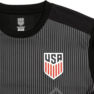 Puede incluir: Camiseta de f&uacute;tbol a rayas negras y grises con un logotipo de EE. UU. rojo, blanco y azul en la parte delantera. La camiseta tiene un cuello negro y mangas cortas.