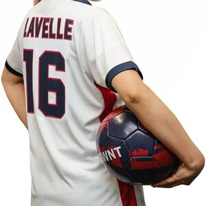 May include: A white soccer jersey with red and blue trim. The jersey has the name "Lavelle" and the number "16" on the back. A person is holding a soccer ball in their right hand.