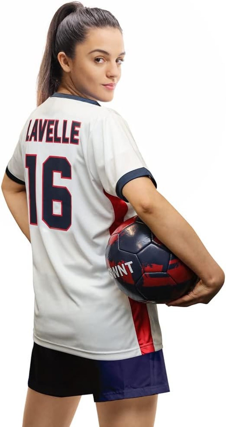 May include: A white soccer jersey with red and blue trim. The jersey has the name "Lavelle" and the number "16" on the back. A person is holding a soccer ball in their right hand.