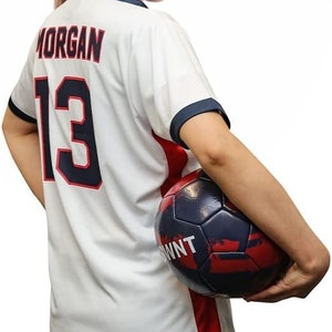 May include: A woman wearing a white football shirt with the name "MORGAN" and the number "13" on the back. She is holding a football in her right hand. The ball is black and red with white lettering.