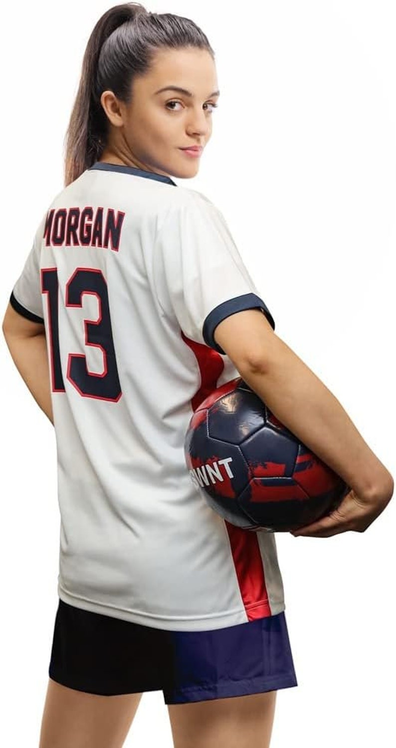 May include: A woman wearing a white football shirt with the name "MORGAN" and the number "13" on the back. She is holding a football in her right hand. The ball is black and red with white lettering.