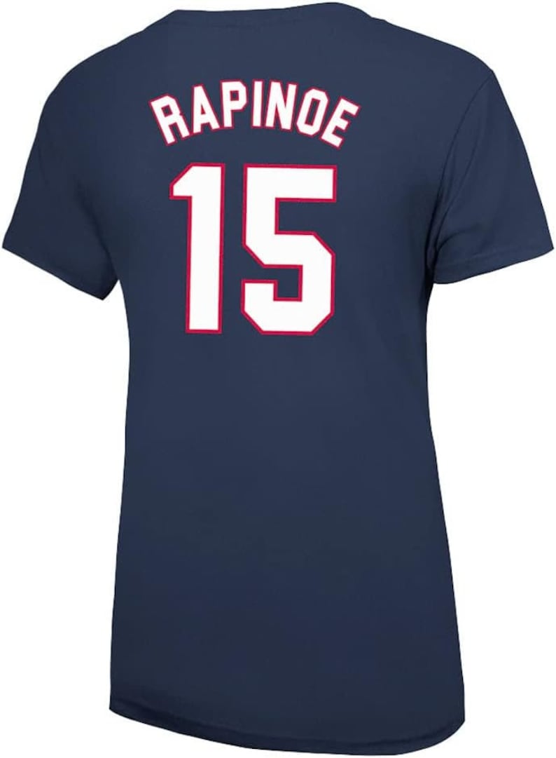 May include: Navy blue t-shirt with the name "RAPINOE" and the number "15" in white with red trim on the back. The shirt is a solid color and appears to be made of a soft material.