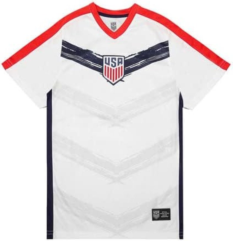 May include: White soccer jersey with red and navy blue accents. The jersey features a stylized USA logo with red, white, and blue stripes. The jersey has a V-neck and short sleeves.