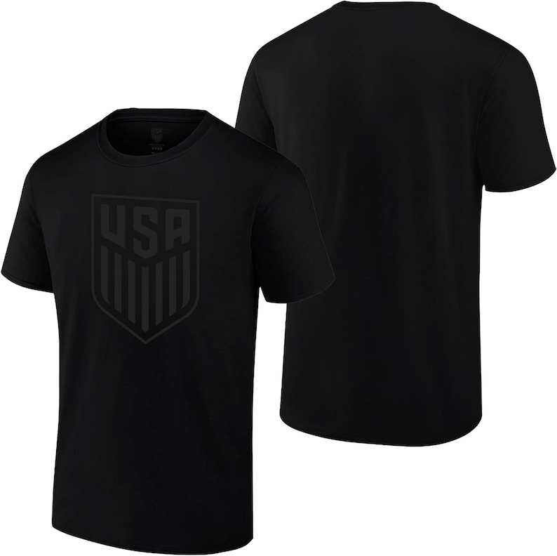 May include: Two black t-shirts. The front shirt features a tonal shield design with the letters "USA" at the top. The shield has vertical stripes. The second shirt is plain black.