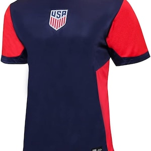 May include: A navy blue soccer jersey with red accents. The jersey has the word "USA" and a shield with red and white stripes on the front.