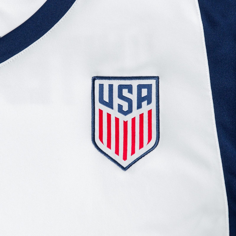 May include: White soccer jersey with a blue and red USA emblem on the chest.