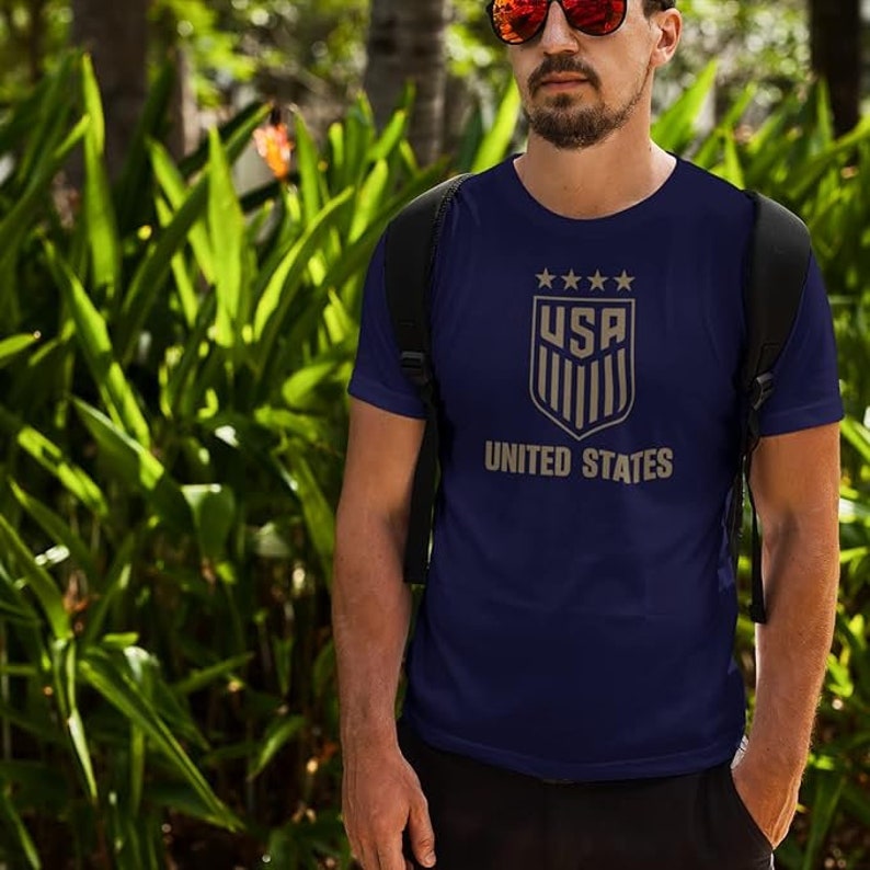 Puede incluir: Una camiseta azul marino con un gr&aacute;fico de escudo dorado que presenta el texto "USA" y "UNITED STATES".