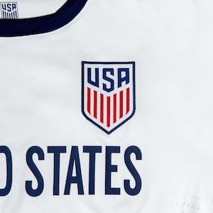 May include: White soccer jersey with a blue and red USA crest and the text "UNITED STATES" in blue.