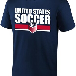 May include: A navy blue t-shirt featuring the text "UNITED STATES SOCCER" in white. Below the text is a red stripe and a USA soccer crest. The shirt has a crew neck design.