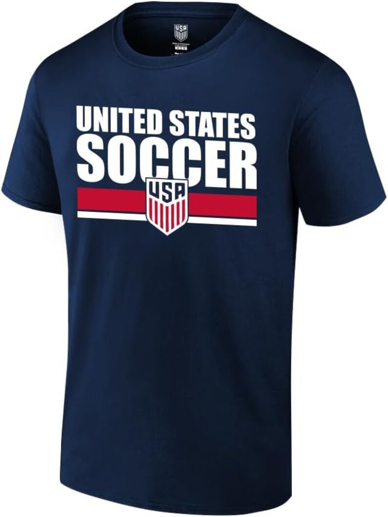 May include: A navy blue t-shirt featuring the text "UNITED STATES SOCCER" in white. Below the text is a red stripe and a USA soccer crest. The shirt has a crew neck design.