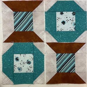 May include: Four fabric squares sewn together to create a larger square. The squares are in a pattern of brown, white, and teal. The teal squares have a white center with a floral pattern and black dots. The brown squares have a white center with a striped pattern in black, white, and teal.