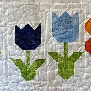 Peut inclure: Un bloc de courtepointe avec une rangée de tulipes bleues et bleu clair avec des tiges et des feuilles vertes. Un seul papillon orange est centré dans la rangée.