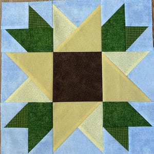 May include: A square quilt block featuring a central brown square surrounded by a yellow starburst design. Green leaf-like shapes are interspersed between the star points, set against a light blue background. The fabrics have various patterns.