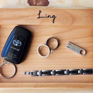May include: A wooden tray with rounded edges, personalised with the name "Ling". The tray contains a car key with a key ring, two silver rings, a silver USB drive labelled "OTG-58 16GB", and a black and silver bracelet.