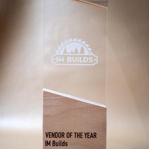 May include: A rectangular award featuring a clear acrylic panel sandwiched between two light-colored wooden blocks. The acrylic displays a white logo of a circular saw blade and trees above the text IM BUILDS. The base reads VENDOR OF THE YEAR IM Builds 2026.