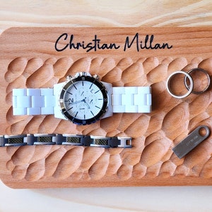 May include: A wooden tray with the name "Christian Millan" engraved on it. The tray has a carved design and holds a white watch, a silver bracelet, two silver rings, and a silver USB drive.