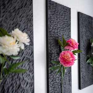 Op de afbeelding: Drie donkergrijze, getextureerde wandpanelen met levendige roze en witte bloemen. De panelen zijn rechthoekig en aan een witte muur gemonteerd, waardoor een moderne bloemenpresentatie ontstaat. De bloemen voegen kleur toe.