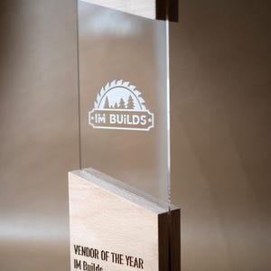 May include: A vertical award featuring a clear acrylic panel sandwiched between two light-colored wooden blocks. The acrylic displays a white circular saw blade logo with trees and the text IM BUILDS. The base is engraved with VENDOR OF THE YEAR IM Builds 2026.