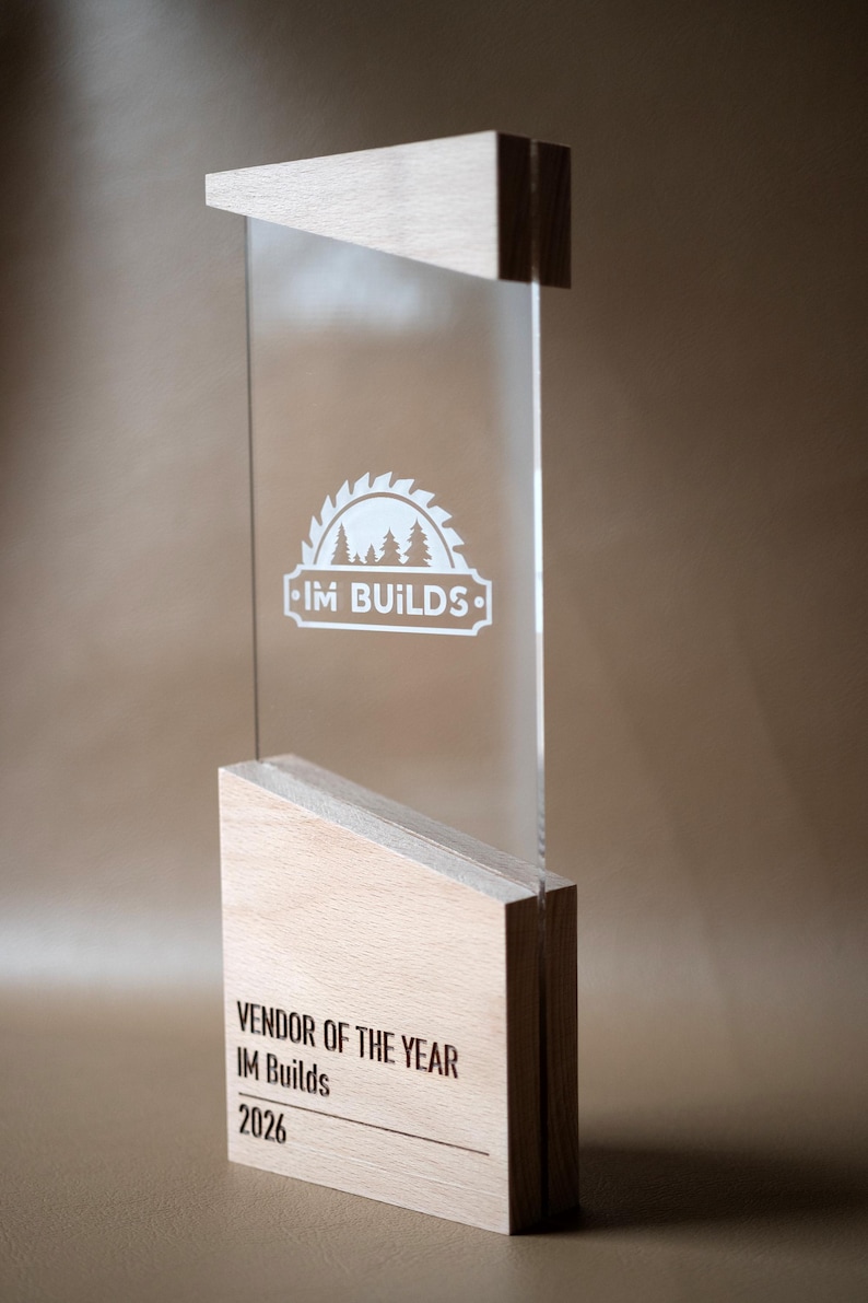 May include: A vertical award featuring a clear acrylic panel sandwiched between two light-colored wooden blocks. The acrylic displays a white circular saw blade logo with trees and the text IM BUILDS. The base is engraved with VENDOR OF THE YEAR IM Builds 2026.