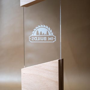 May include: A clear acrylic rectangular plaque held between two light-colored wooden blocks with angled tops. The acrylic features a white etched graphic of a circular saw blade containing pine trees and the text "IW BUILDS".