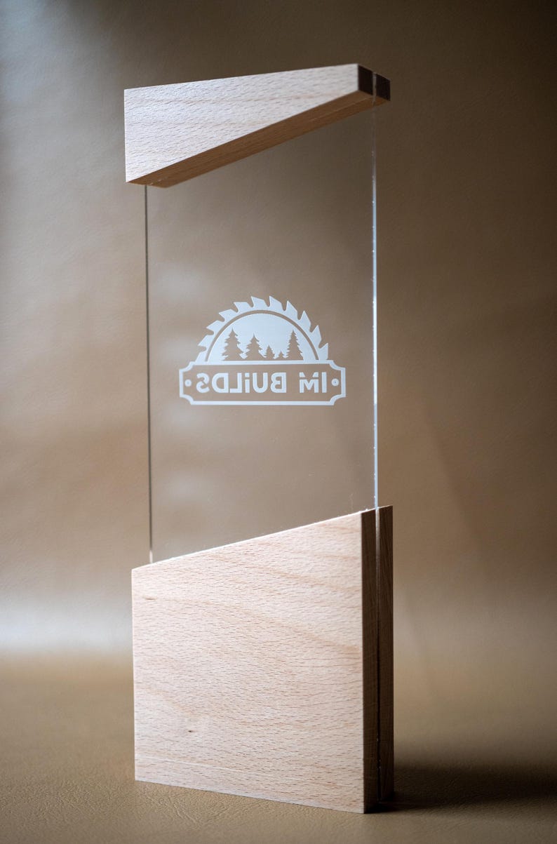 May include: A clear acrylic rectangular plaque held between two light-colored wooden blocks with angled tops. The acrylic features a white etched graphic of a circular saw blade containing pine trees and the text "IW BUILDS".