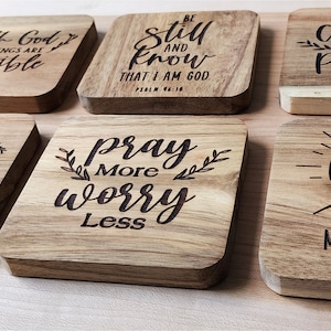 May include: Eight wooden coasters with inspirational quotes and designs. The quotes include "With God all things are possible", "I am loved", "Be still and know that I am God", "Pray more worry less", "Created with a purpose", "Faith can move mountains", and "Matthew 17:20".