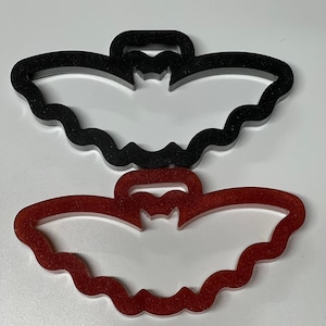 May include: Two bat-shaped cookie cutters, one black and one red. The cutters are made of plastic and have a scalloped edge.