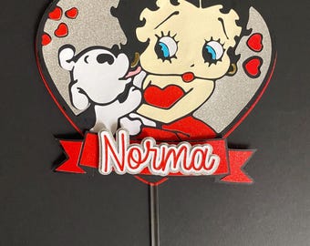 May include: A heart-shaped cake topper featuring a cartoon image of Betty Boop with a white dog. The design includes red hearts and the name "Norma" in a banner. The topper is on a silver stick.