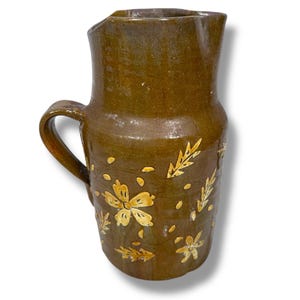 May include: A handcrafted, olive-green ceramic pitcher with a handle and a spout. The pitcher is adorned with hand-painted yellow floral and leaf designs. The rustic pottery piece is suitable for serving beverages or as a decorative item.