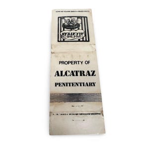 May include: Vintage matchbook from Alcatraz Penitentiary. The matchbook is off-white with black text that reads "Property of Alcatraz Penitentiary." The top section features a graphic and the text "Spent Time on Alcatraz."
