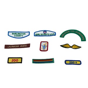 May include: A collection of embroidered patches in various shapes and colors. Includes patches with text like "Burlington County," "Girl Scouts USA," and "Junior Aide." Other patches feature a jumping rope design, a pair of wings, and the numbers "200" and "200+."