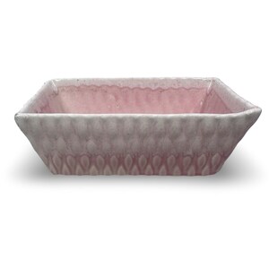May include: A rectangular ceramic dish with a light pink interior and a textured, light gray exterior. The exterior features a repeating pattern of small, oval shapes. The dish has a slightly flared rim and is likely used for serving or display.