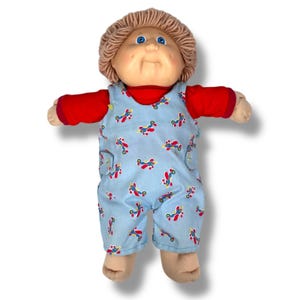 May include: A vintage doll with a soft, fabric body. The doll has a round face, blue eyes, and light brown yarn hair. It wears a red long-sleeved shirt and light blue overalls patterned with red and blue airplanes. The doll's arms are outstretched.