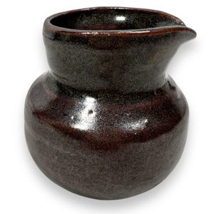 May include: A small, dark brown ceramic pitcher with a rounded body and a pouring spout. The pitcher has a glossy finish with subtle variations in color, ranging from deep brown to hints of green. The rim is a darker shade of brown.