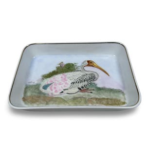 May include: Rectangular ceramic tray with a painted illustration of a white ibis bird with pink accents, a yellow beak, and a gold rim. The bird is perched on a green and brown landscape against a light blue background.