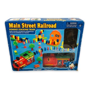 May include: A boxed Main Street Railroad Domino Stacking Train set. The box is blue with images of the train, buildings, and dominoes. The train is red and green, with the number 1 on the side. The box also features the Disneyland and Walt Disney World logos.