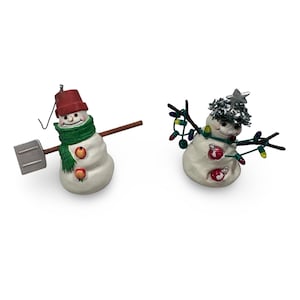 May include: Two snowman ornaments. One snowman wears a red bucket hat, green scarf, and holds a shovel. The other snowman has a silver star on its head and is decorated with Christmas lights and ornaments.
